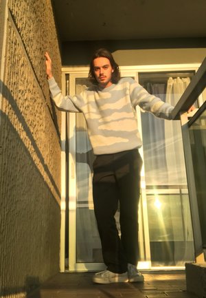 Suéter azul claro con patrón de nubes, pantalones negros y zapatos blancos. De pie en un balcón con pared texturizada y barandilla de cristal.