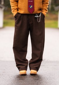 Personne portant un pantalon marron à jambes larges, des bottes jaunes, un pull orange et une cravate rouge avec un motif de cœur et de pansement.