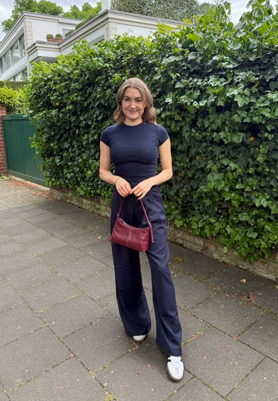 Top ajustado de color navy combinado con pantalones de pierna ancha también en navy. Sosteniendo un pequeño bolso burdeos. Zapatillas casuales. Follaje verde en el fondo.