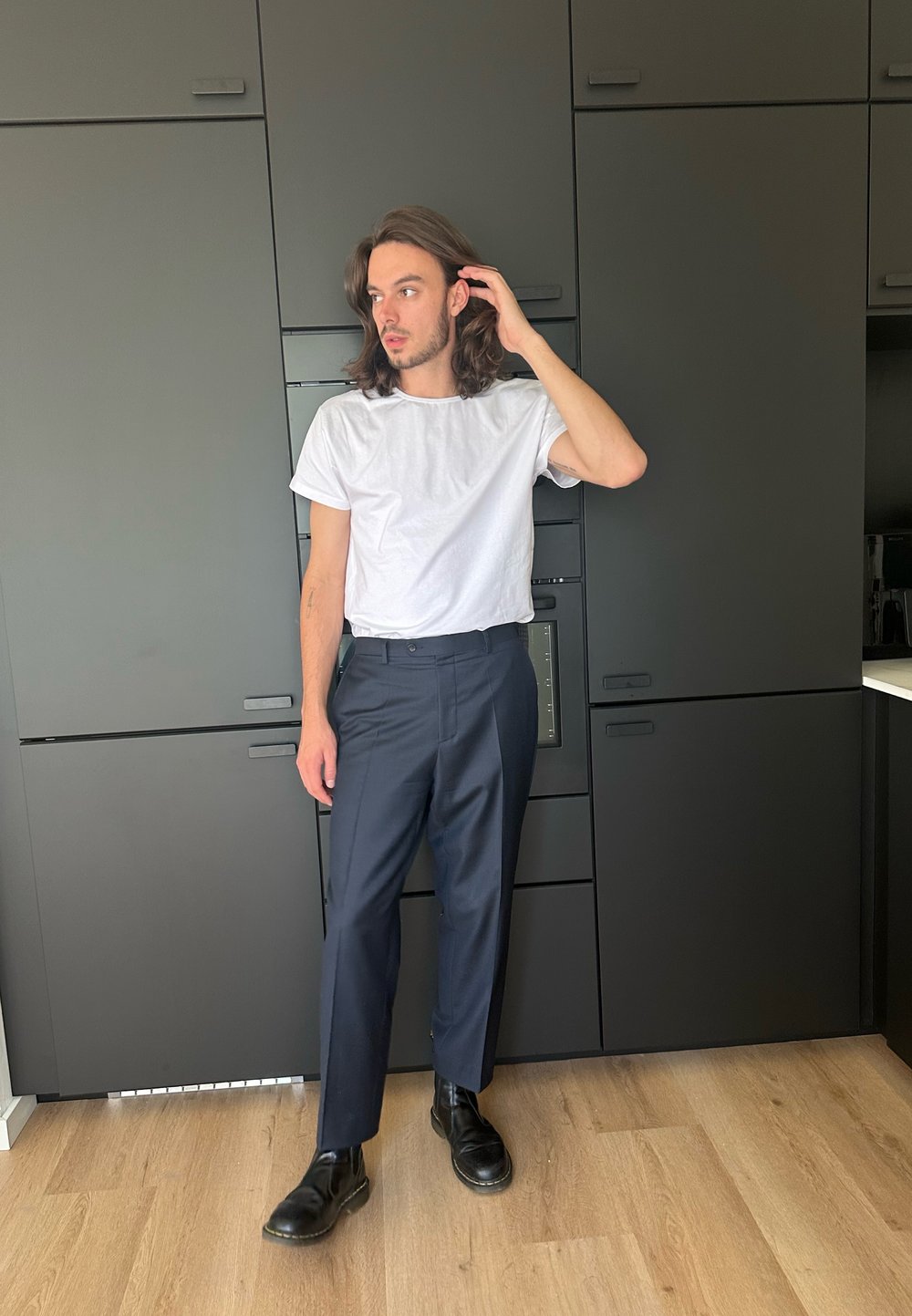 Camiseta branca de gola redonda, calças ajustadas azul-marinho e botas de couro pretas. Cabelo longo e pose casual contra um armário escuro com chão de madeira.