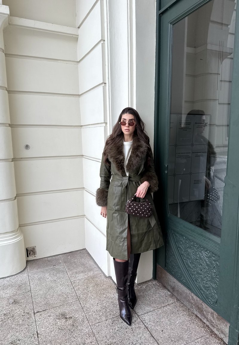 Femme portant un long manteau vert à col en fourrure, bottes noires montantes jusqu'aux genoux, lunettes de soleil, tenant un petit sac à main clouté, debout près d'une porte verte.