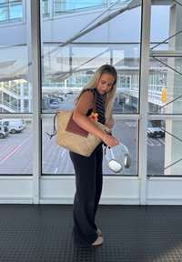 Torebka tote z plecionego słomy z brązowym skórzanym akcentem, zdobiona detalem w kształcie kwiatu. Użytkownik trzyma srebrne słuchawki, ubrany w czarne szerokie spodnie.