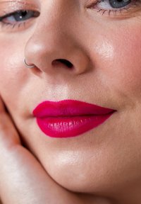 Heldere fuchsia lippenstift op gladde, goed gedefinieerde lippen. Een zilveren neusring accentueert het gezicht van het model. De huidtextuur is zacht en egaal.