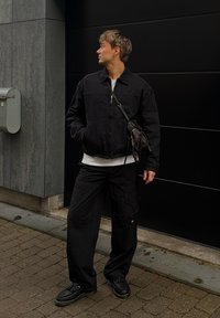 Čierna bomber bunda cez bielu košeľu, v kombinácii s voľnými čiernymi cargo nohavicami a čiernymi topánkami. Malá taška nosená cez telo, vonkajšie prostredie.