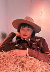 Donna con capelli scuri, che indossa un cappello beige a tesa larga e un giacca marrone con spille, sdraiata su un mucchio di paglia all'interno con sfondo neutro.