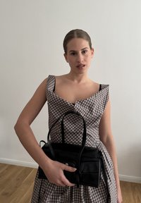 Jeune femme aux cheveux tirés en arrière portant une robe sans manches à carreaux, tenant un sac à main en cuir noir contre un mur blanc uni.