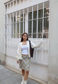 Bílá bomber bunda přes bílou blůzu, vzorovaná sukně s zelenými a modrými kosočtverci, hnědá pletená taška, béžové kotníkové boty.