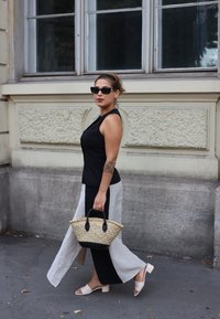 Femeie care merge pe strada din oraș, îmbrăcată cu un top negru fără mâneci, pantaloni bifocali alb-negru, sandale cu toc bej, ochelari de soare și purtând o geantă împletită.