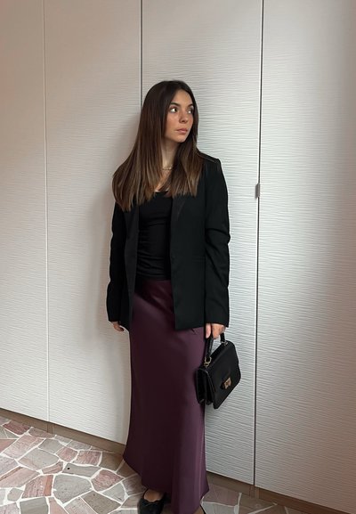 Blazer negro sobre una blusa negra, combinado con una falda larga y lisa de color burdeos. Lleva un pequeño bolso negro. La pared presenta un diseño texturizado.