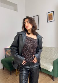 Černá kožená bunda přes top s leopardím potiskem, doplněná lesklými černými umělými koženými kalhotami. Pozadí tvoří zelená pohovka a rámované umělecké dílo.
