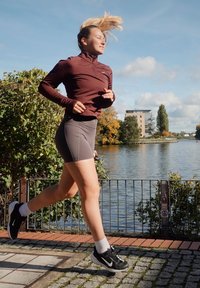 Top com fecho éclair na cor bordô, calções cinzentos e sapatilhas de corrida pretas. A correr ao lado da água, com vegetação e edifícios ao fundo.