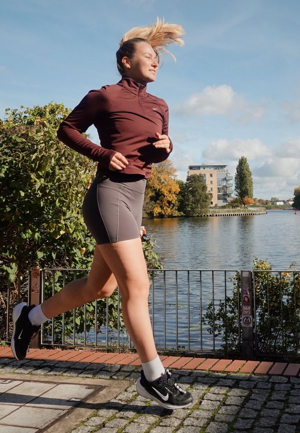 Haut zippé bordeaux, shorts gris et chaussures de course noires. En train de courir à côté de l'eau avec de la verdure et des bâtiments en arrière-plan.
