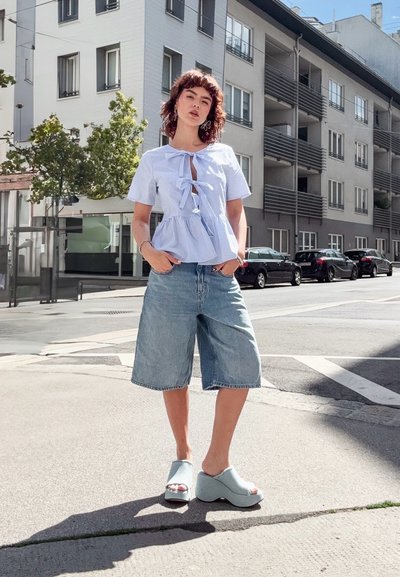 Top peplum a rayas azul claro con detalle de lazo, combinado con pantalones cortos de denim sueltos y sandalias de cuña en azul pálido, con un fondo urbano.