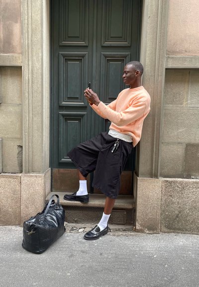 Una persona está de pie junto a una puerta de madera verde, vistiendo un suéter naranja, pantalones cortos negros y mocasines negros combinados con calcetines blancos. Hay una bolsa negra junto a ellos.