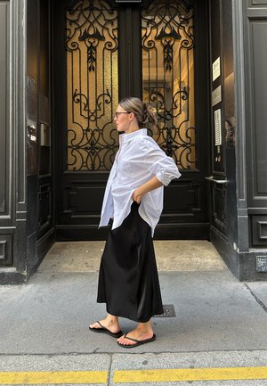 Camisa blanca oversize, falda maxi de satén negra y chanclas negras. Puerta oscura y ornamentada con paneles de vidrio de fondo.