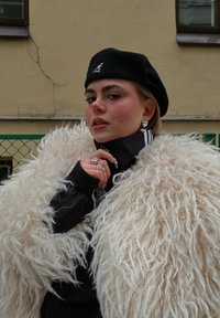 Model wearing a fluffy white fur coat, a black jacket with white stripes, and a black beret. Notable jewellery includes heart-shaped earrings and multiple rings.
