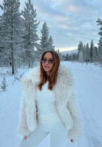 Vit fuskpälsjacka med en yvig textur, bärs över en figurnära vit topp och leggings, mot en snötäckt skogsbackgrund. Klara solglasögondetaljer.