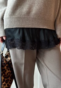 Béžový pletený svetr přehozený přes černou košili s krajkovým lemem, doplněný šedými kalhotami. Na černém popruhu visí taška se vzorem leopardí kůže.