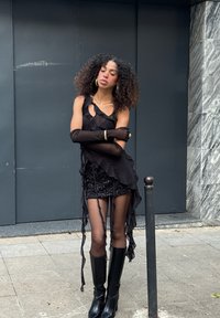 Schwarzes, mehrlagiges Oberteil mit Volants, kombiniert mit einem schimmernden Minirock. Lange schwarze Handschuhe und kniehohe Stiefel mit Absatz vervollständigen das Outfit.