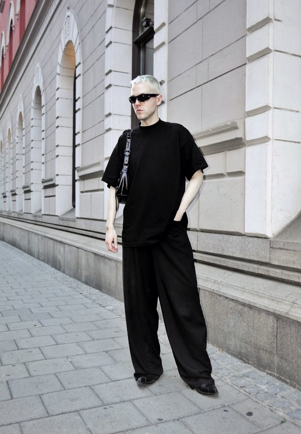 Osoba s krátkymi blond vlasmi, oblečená v čiernom voľnom oblečení a slnečných okuliaroch, stojí na chodníku pri kamennej fasáde budovy.