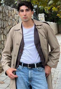 Beige Trenchcoat über einem braunen Cardigan, weißem T-Shirt und heller blauer Jeans. Verfügt über graue Knöpfe und einen schwarzen Gürtel mit silberner Schnalle.