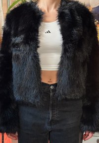 Jachetă scurtă din blană artificială neagră cu finisaj texturat, cu deschidere frontală și mâneci lungi, purtată peste un maiou alb și blugi negri.