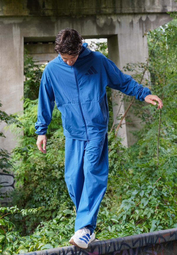 Man in blue tracksuit balancing on a graffiti-covered metal rail surrounded by green plants under a concrete bridge.