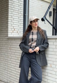 Tumedat halli tooniga laia säärega püksid, mida on rõhutatud ruudulise halli bleiseri ja beežika mustriga kampsuniga. Komplekti lõpetab hele nokkmüts. Lihtne taust.