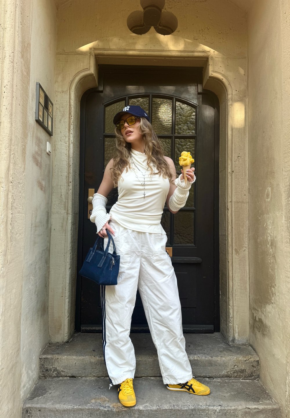 Top bianco senza maniche, pantaloni bianchi larghi con strisce nere, sneakers gialle, cappellino blu scuro, occhiali da sole, in mano un gelato, piccola borsa blu.