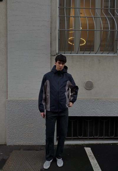 Joven con chaqueta negra y gris y zapatos a cuadros de pie sobre un pavimento oscuro frente a una ventana con rejas en una pared gris.
