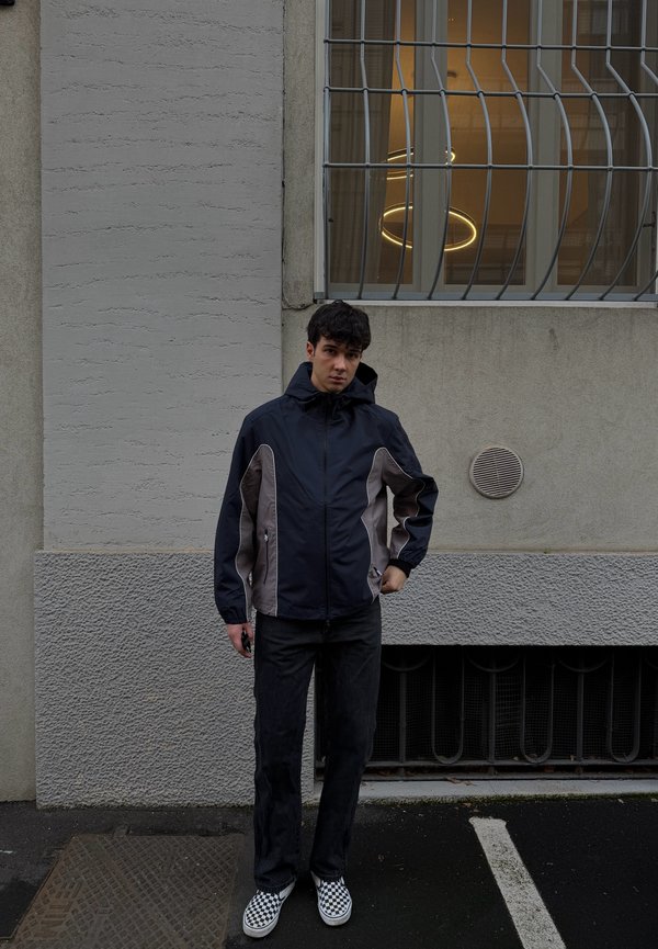 Fiatal férfi fekete és szürke dzsekiben, kockás cipőben áll egy sötét járdán egy szürke falon lévő rácsos ablak előtt.