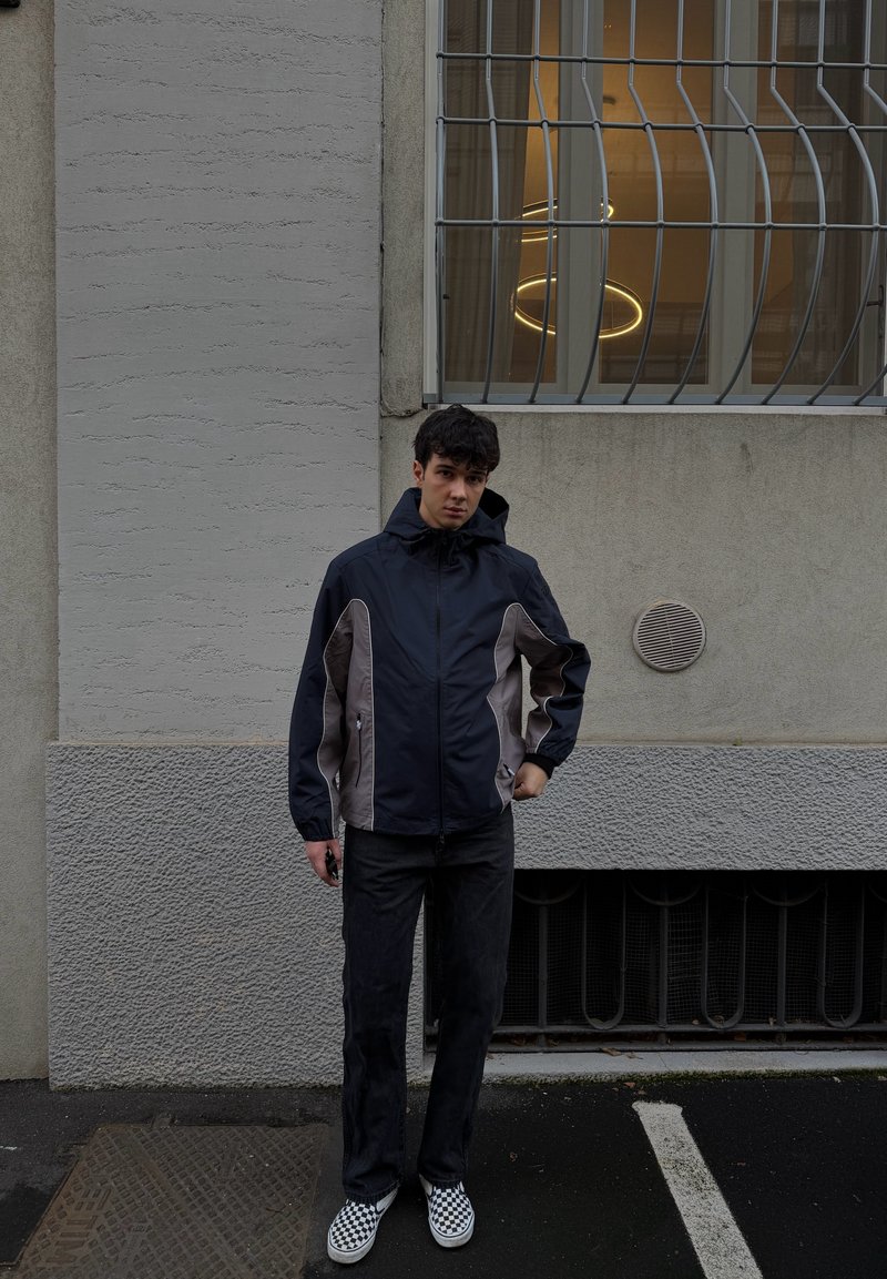 Mladý muž v čierno-šedej bunde a kockovaných topánkach stojí na tmavom povrchu pred oknom so železnými mrežami na šedej stene.