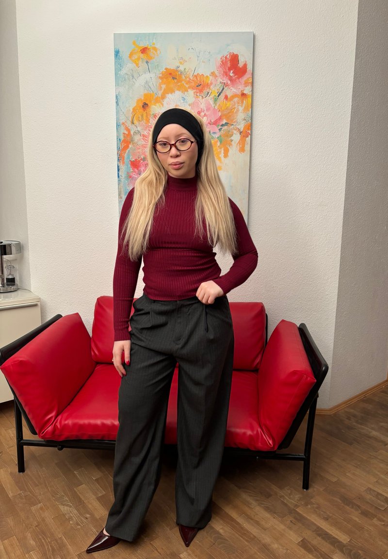 Maroon ribbed turtleneck, dark pinstriped wide-leg pants, and burgundy pointed heels. Red couch in background and colorful floral artwork.