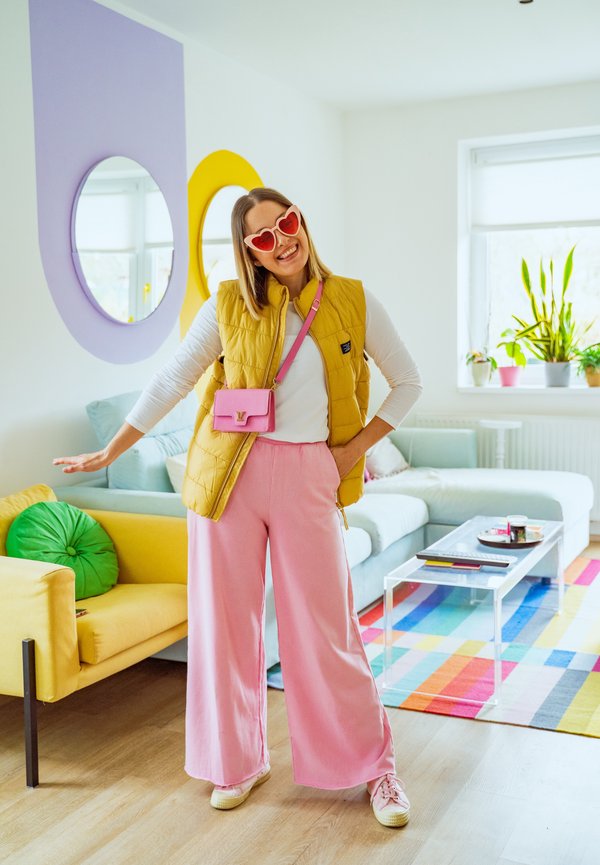 Kvinne med hjerteformede solbriller, gul vest og rosa bukser som står i en fargerik stue med moderne møbler og planter ved et vindu.