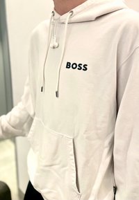 Felpa bianca con tasca frontale, logo "BOSS" nero, coulisse e cuffie appese al collo. Tessuto morbido e vestibilità rilassata.