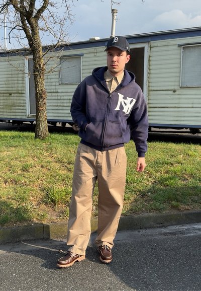 Sudadera de marina con logo blanco de NY, pantalones anchos beiges y zapatos marrones con cordones y suelas texturizadas. El fondo incluye un área de hierba verde.