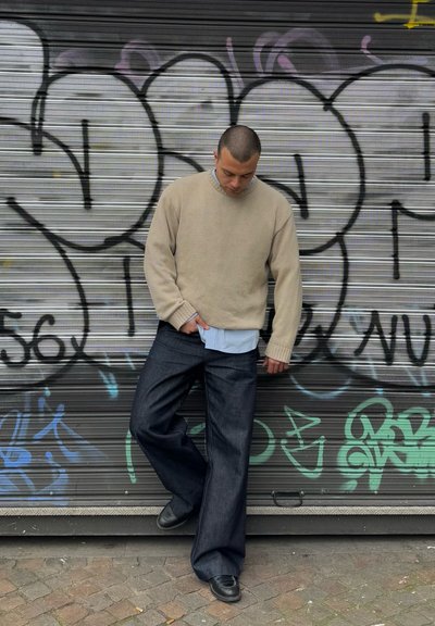 Suéter beige texturizado, camisa azul clara debajo y pantalones vaqueros de pierna ancha azul oscuro. De pie contra una pared cubierta de graffiti. Zapatos negros.