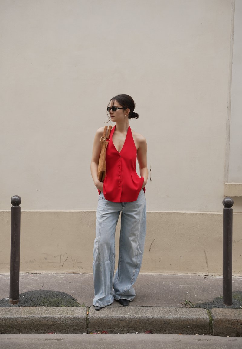 Femme portant un haut rouge sans manches à boutons, un jean large bleu clair, des chaussures noires, des lunettes de soleil, et portant un sac à bandoulière marron, debout sur un trottoir.
