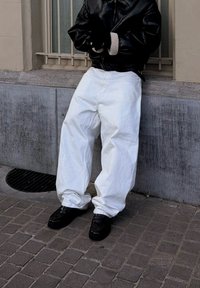 Personne portant un pantalon blanc surdimensionné, des chaussures noires et une veste en cuir noire avec des poignets en mouton, debout sur un pavé en pierre.