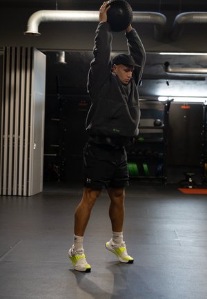 A person in a dark gray fleece top and black shorts lifts a textured black medicine ball overhead. They wear white and neon yellow athletic shoes.