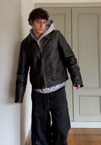 Jeune homme portant une veste en cuir noire par-dessus un sweat à capuche gris et un jean noir ample, debout à l'intérieur contre un mur clair et des portes fermées.