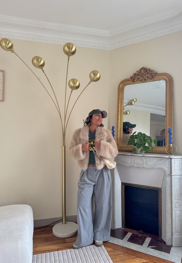Ung person i afslappet tøj og pelsjakke står ved en vintage marmorkamin med et guldrammet spejl og en gulvlampe i messing.