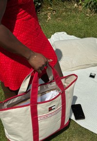 Bolsa de lona beige con detalles y asas rojas, que presenta el logo de "TOMMY HILFIGER". En el interior contiene papel blanco. Teléfono inteligente negro sobre una manta.