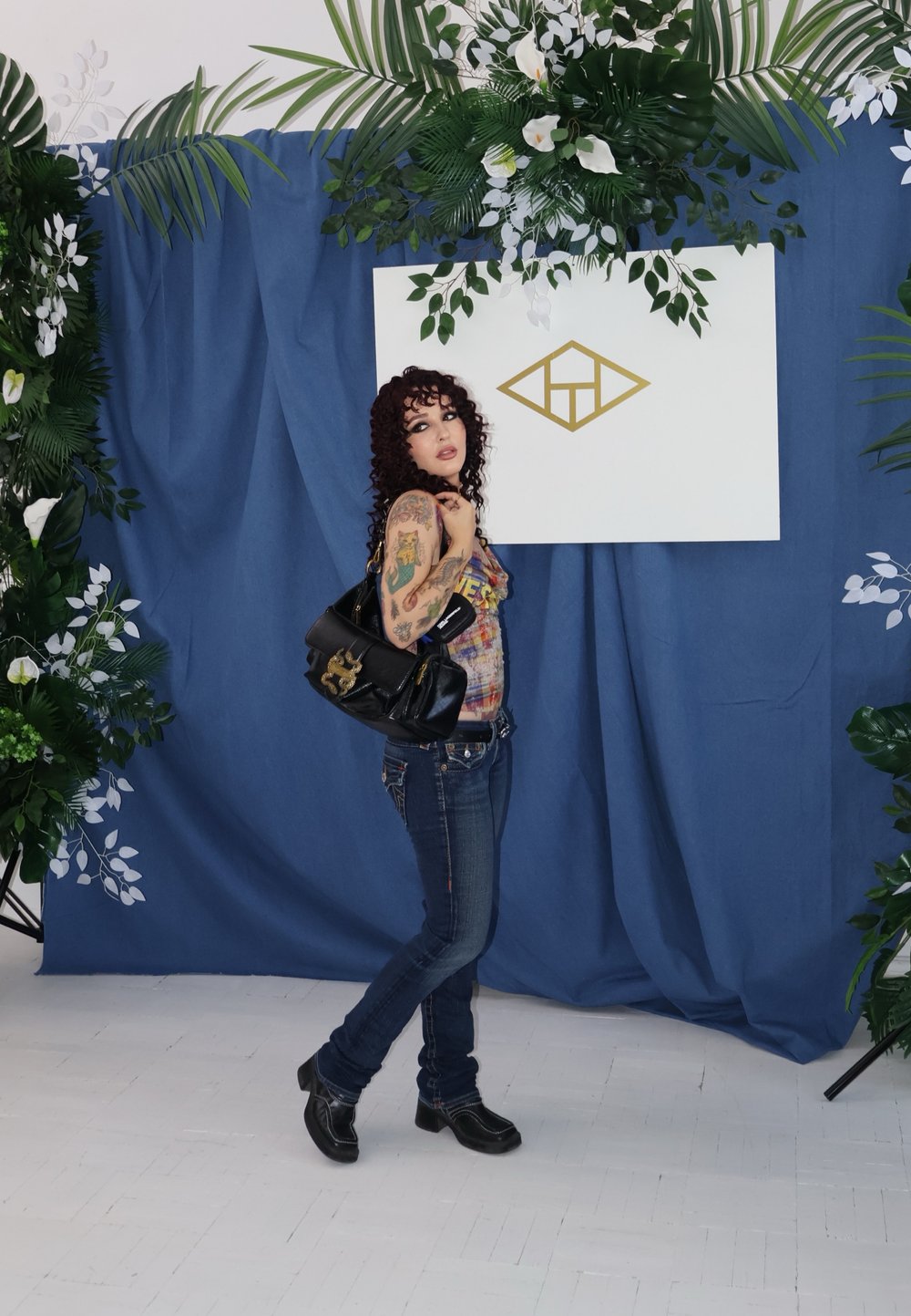 Must musta kott, millel on kuldne ripats, kantud õlal. Mudel, kellel on seljas värviline kampsun ja sinised teksad, seisab sinise tausta ees.