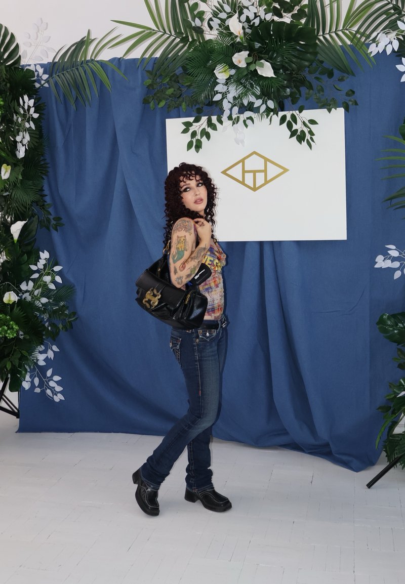 Zwarte leren handtas met een gouden hardware accent, gedragen op de schouder. Model in een kleurrijk tanktop en blauwe jeans staat tegen een blauwe achtergrond.