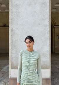 Langermet topp i lys mintgrønn med rynkedetaljer, ribbet tekstur og slim fit, kombinert med et matchende skjørt. Bakgrunnen viser en steinvegg.