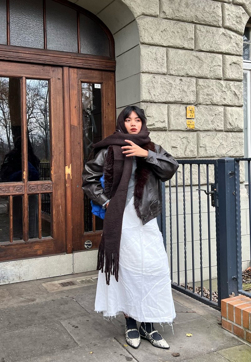 Giacca di pelle marrone sopra una maglia grigia, abito bianco con frange e lunga sciarpa scura. Scarpe nere e argento puntate, in piedi accanto a una porta di legno.