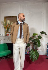 Veste marron zippée avec col contrastant, chemise bleu clair, cravate tricotée marron et pantalon large crème. Cadre intérieur avec des plantes vertes.