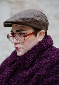 Personne portant une casquette plate marron, de grandes lunettes à monture marron, plusieurs piercings aux oreilles, un anneau au septum et une écharpe texturée violette.