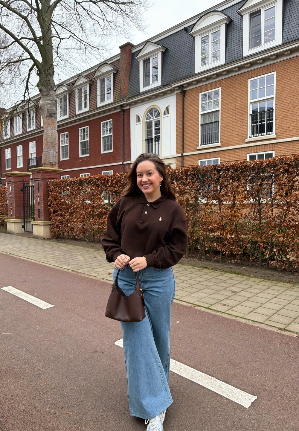 Jeune femme souriante tenant un sac à main marron, debout dans une rue avec des bâtiments en briques et des arbres sans feuilles en arrière-plan.